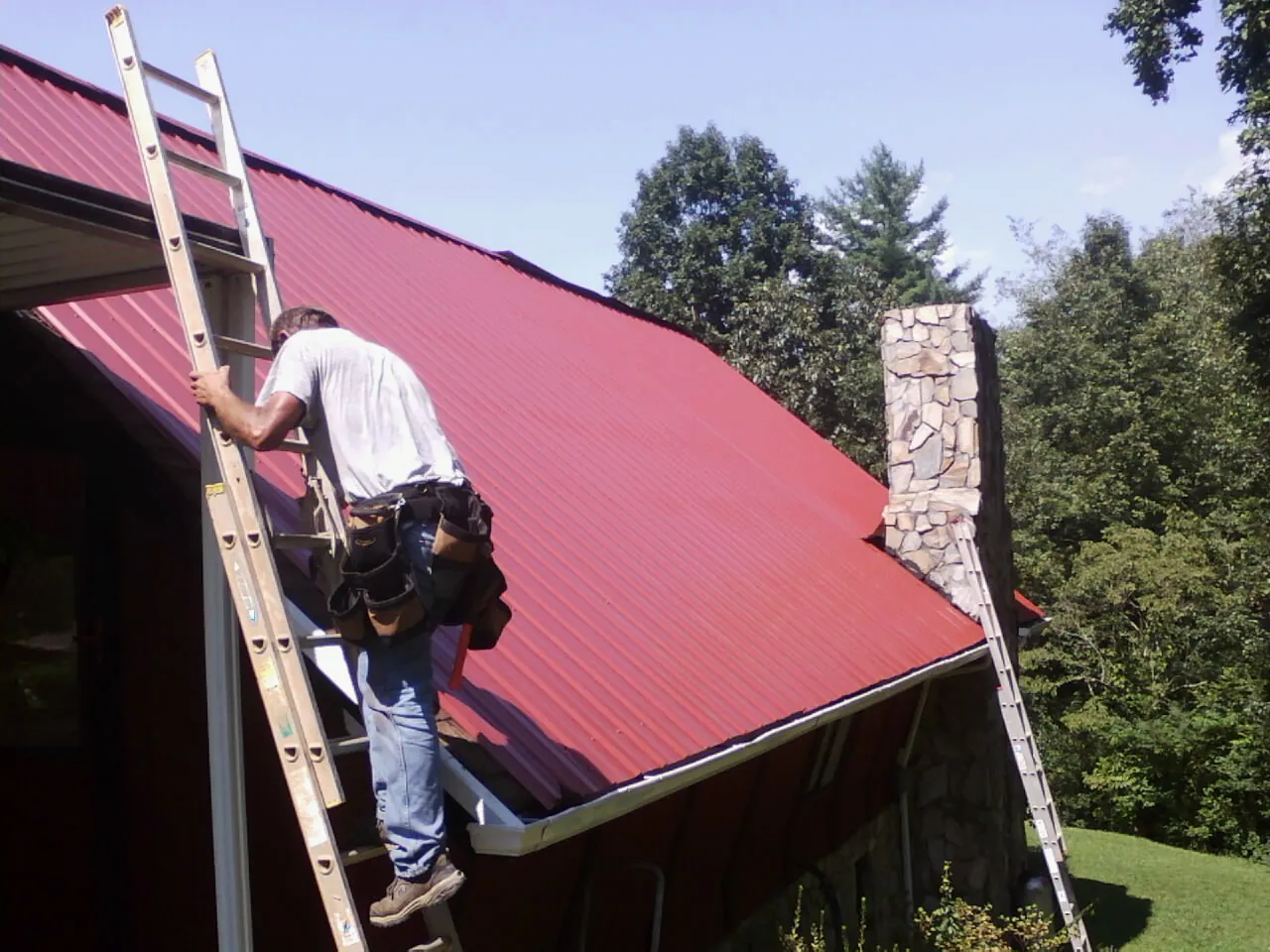Roofer on a ladder beside a red metal roof and stone chimney — Soffit & Fascia Repair by Roofing Brothers in West Plains